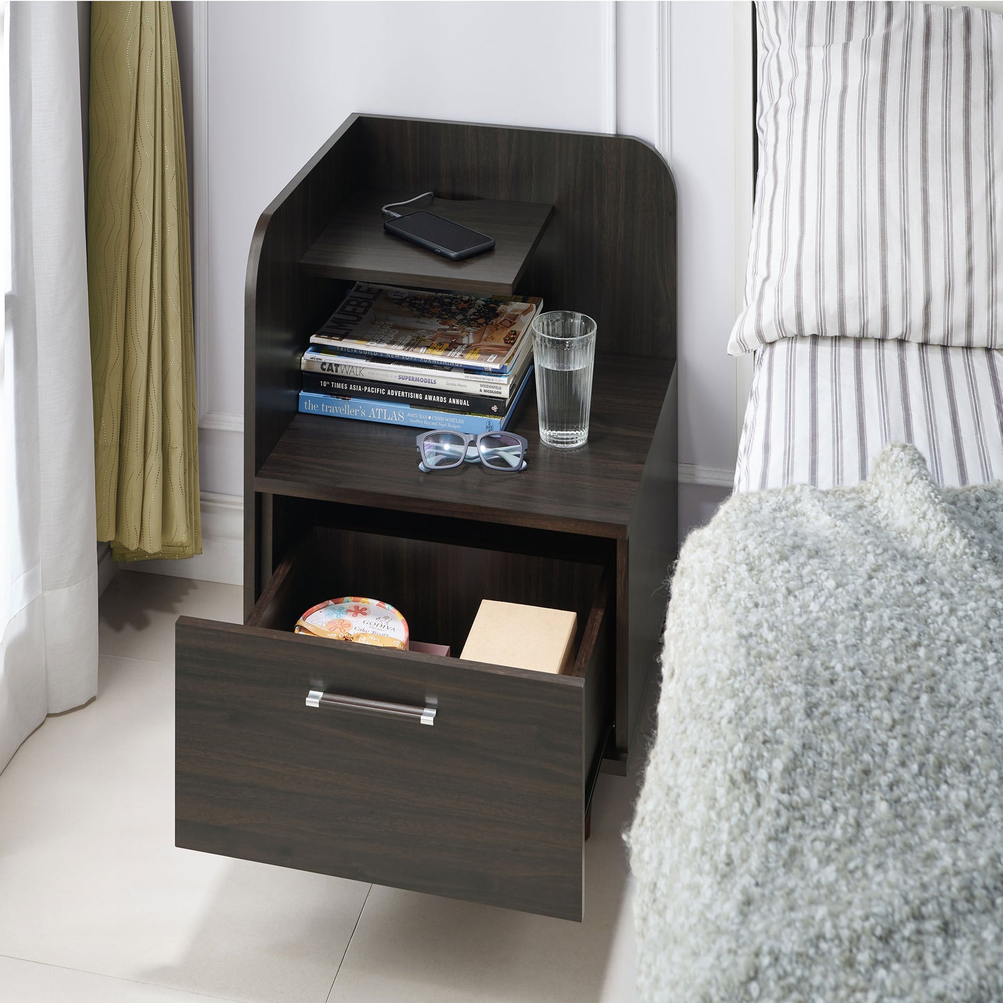 Left angled upper view of a contemporary wenge one-drawer nightstand with a mini shelf and drawer open in a bedroom with accessories