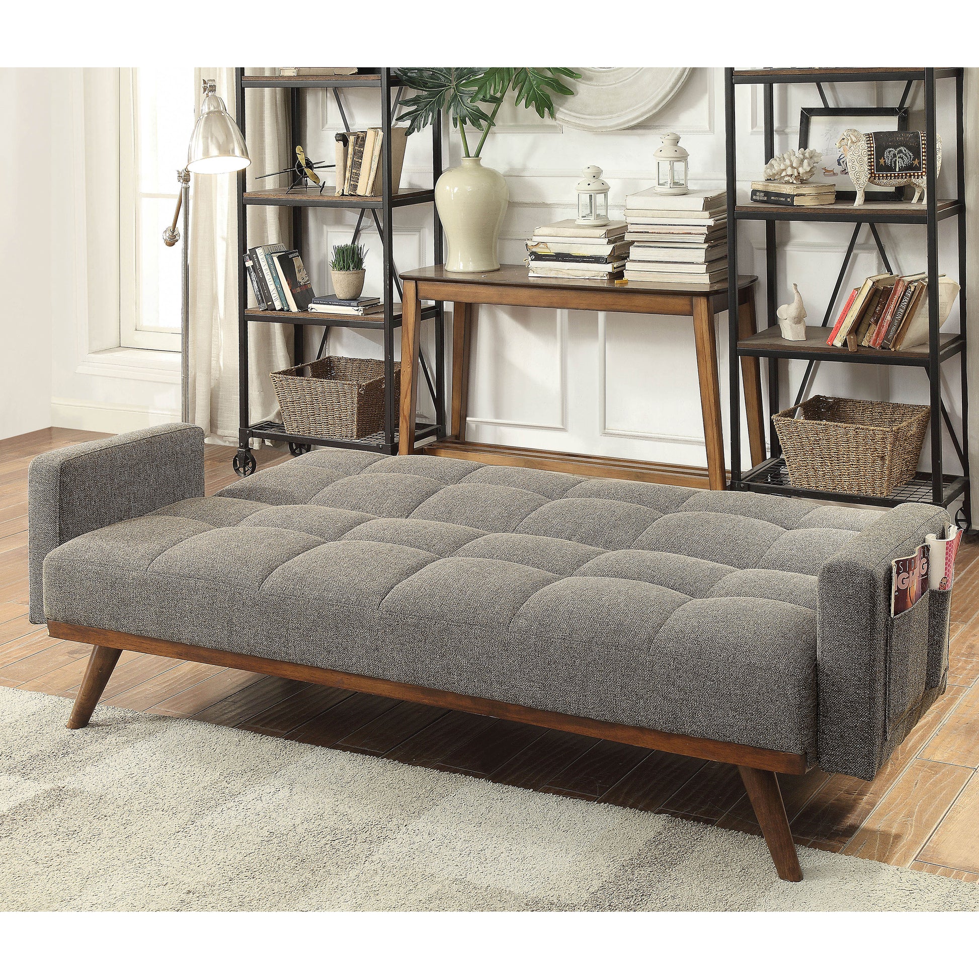 Left angled mid-century modern gray tufted futon with a wood base laid flat in a living area with accessories