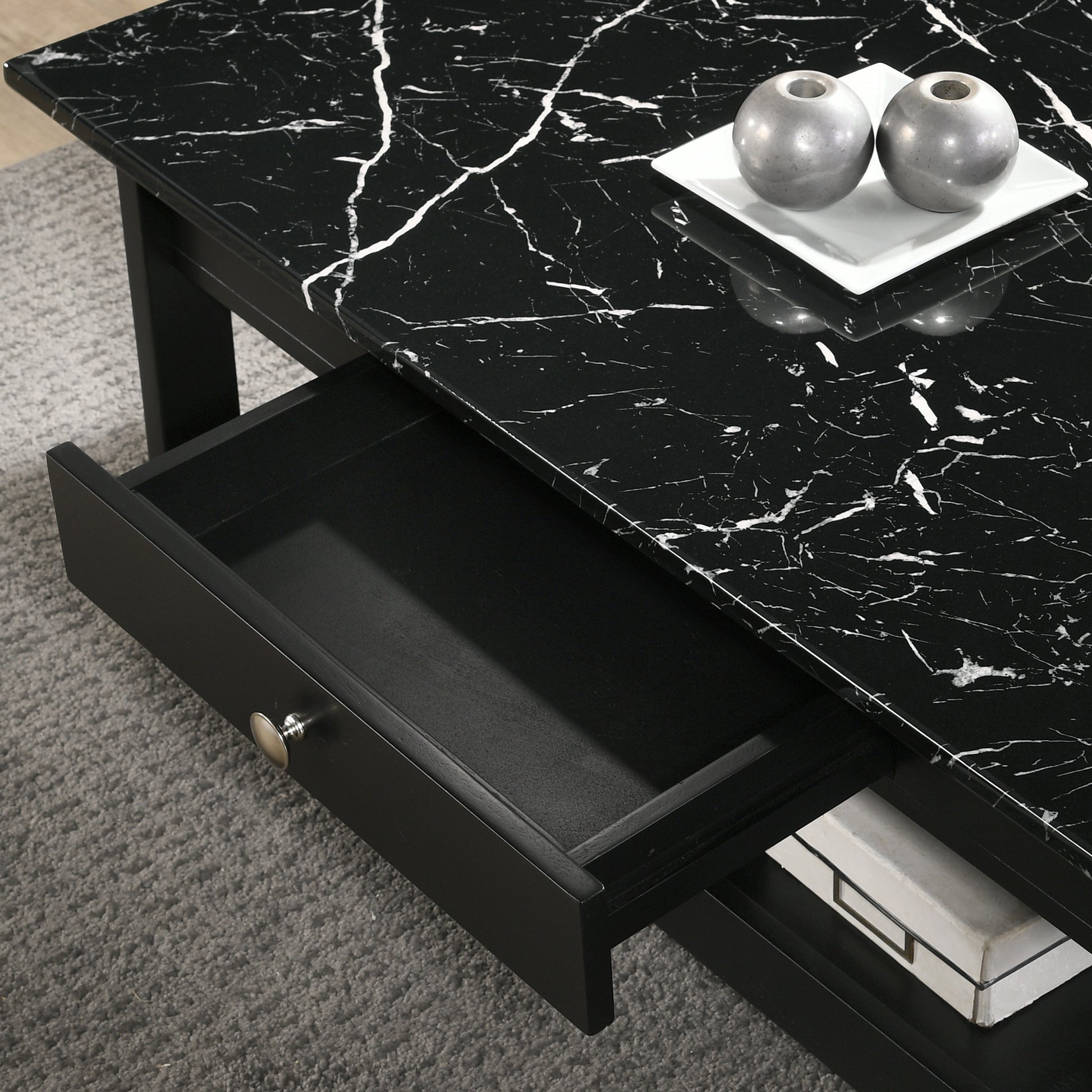 Left angled coffee table only from a three-piece modern black and faux marble coffee table set with lower shelves and drawer open in a living area with accessories